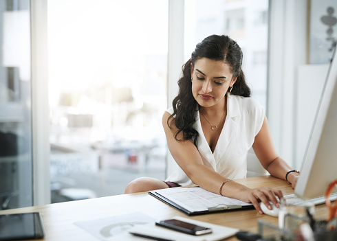 Shot of a young businesswoman using a computer and going through paperwork at her desk in a modern office