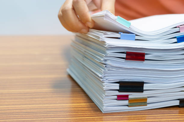 istock Teacher hand searching report papers and students homework assignment for checking score. Pile of unfinished paperwork stacked in archive with colorful paper clips. Business and education concept. 1165989029