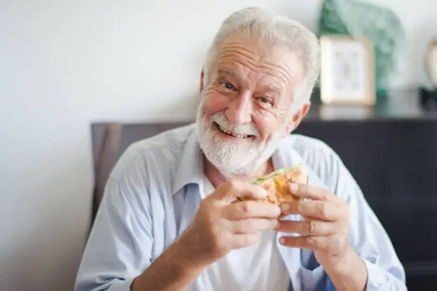 Photo of Seniors European man is sitting to eat a burger at home which looking at camera. Retired man is smiling with felling happy and take a burger on hand.