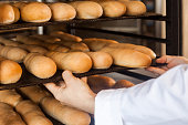 Closeup of hands of professional chef man in white uniform standing near shelves full with fresh bread and pulling baking tray for checking quality
