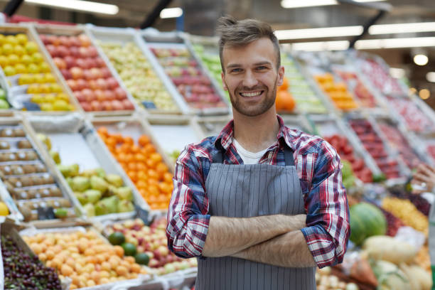 Man Working in Supermarket Waist up portrait of handsome young man working in supermarket and smiling at camera while posing by fruit stand, copy space market vendor photos stock pictures, royalty-free photos & images
