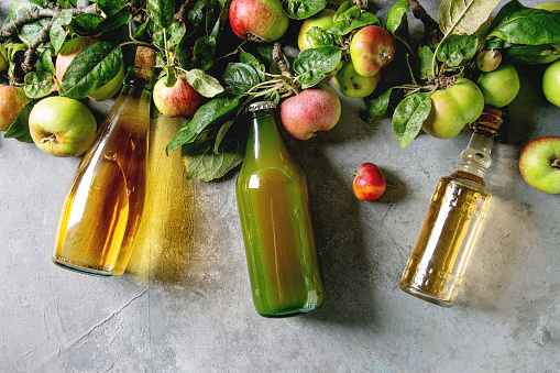 Variety of apple drinks. Bottles of apple juice, vinegar and cider with garden apples with leaves and branches over grey texture background. Flat lay, space. Autumn home harvesting.