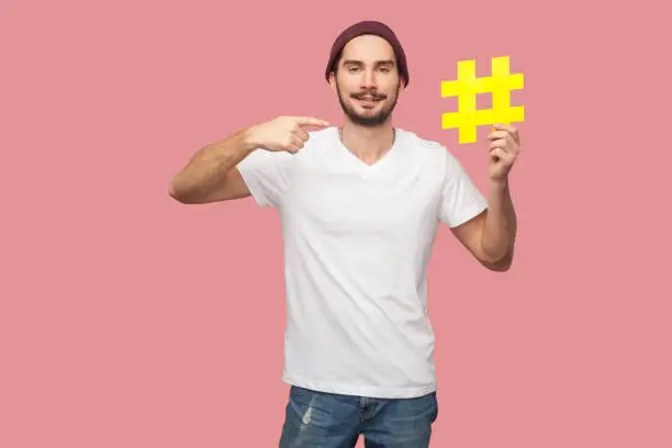Photo of Portrait of happy handsome bearded young hipster man in white shirt and casual hat standing, holding yellow hashtag and pointing finger to it.