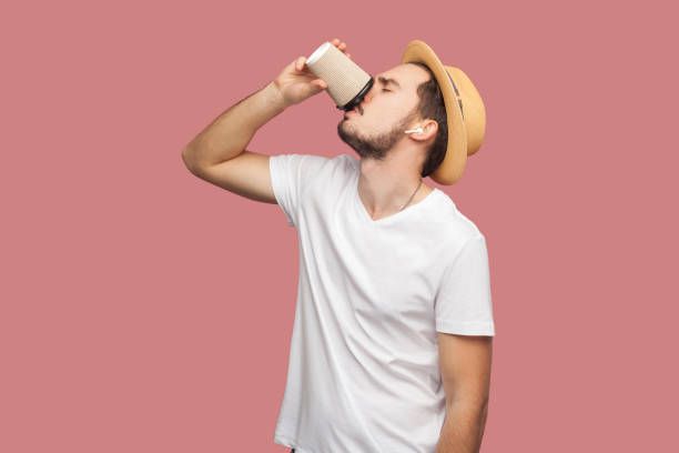 verticale de l'homme jeune barbu beau dans la chemise blanche et le chapeau occasionnel restant et buvant le café dans la tasse en papier, mode de vie de fraîcheur. - fresh coffee audio photos et images de collection