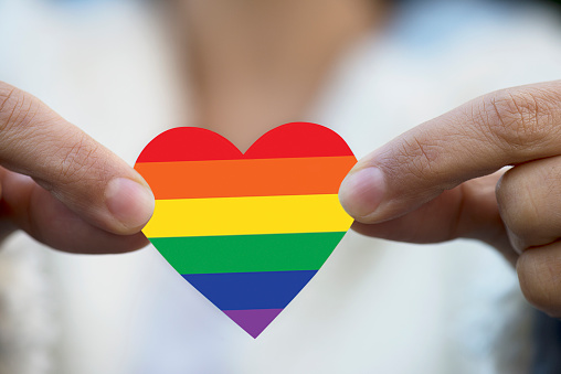Fingers are showing a heart shape to camera made of rainbow flag colors symbolizing LGBT movement.