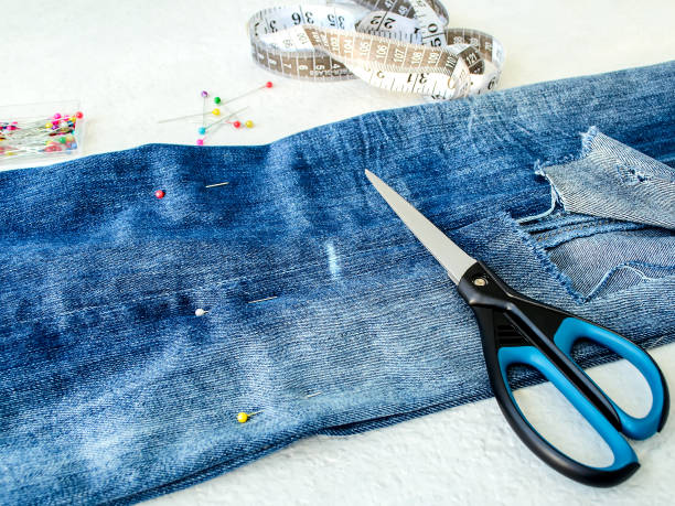 istock Blue jeans with large hole folded in half, sewing pins, white tailor tape with centimeters and inches and scissors. Shorten the jeans. DIY shorts out of jeans. 1166084596