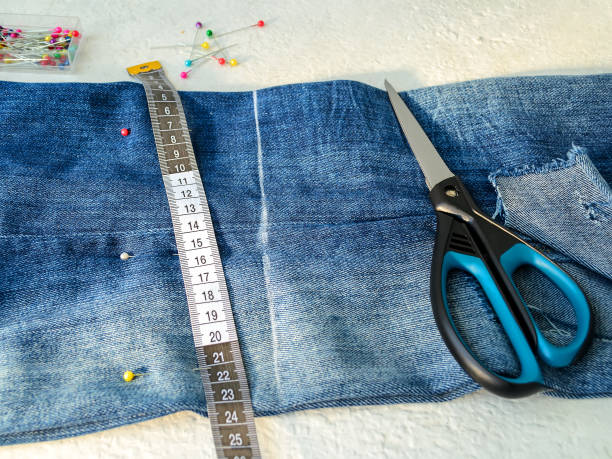 istock Blue jeans with large hole folded in half, multi colored sewing pins in a box, white tailor tape with centimeters and inches and scissors. Shorten the jeans. DIY shorts out of jeans. 1166085633