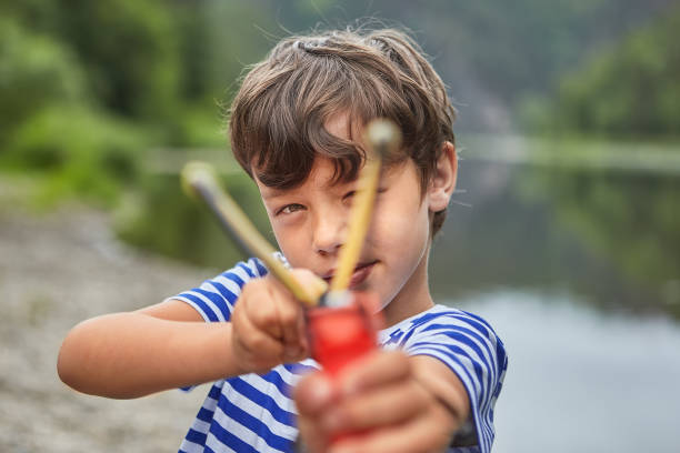 White bully boy about 8 years old is playing with slingshot in park and preparing for shot.