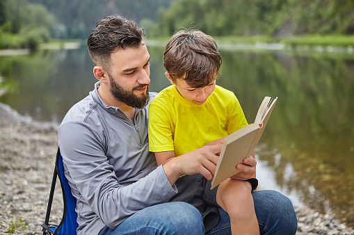 Young attractive caucasian man and his son are sitting on the river bank and reading a book, eco-tourism.