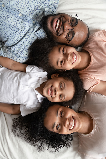 Top view portrait of smiling young african American parents lying in row on cozy bed with happy little boy and girl kids, laughing black family with children look at camera posing relaxing in bedroom