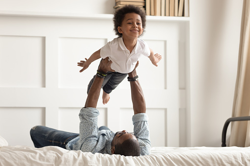 Loving african American dad lying on bed playing with cute little toddler son acting plane, happy black father have fun engaged in funny activity with small kid hold lift in arms in bedroom