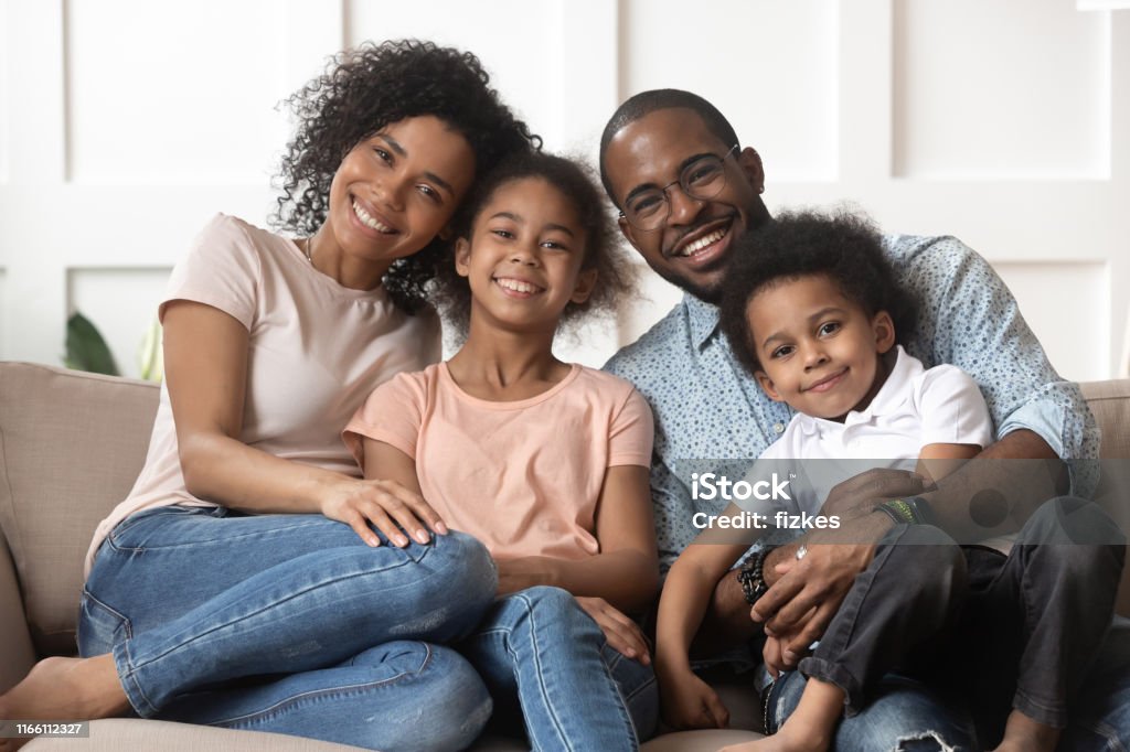 Portrait of black family with kids relax on couch Portrait of happy young african American family with little kids sit relax on couch cuddling, smiling black parents rest on sofa hug preschooler children posing for picture at home together Family Stock Photo