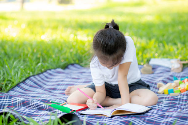 o livro asiático pequeno novo da leitura e da escrita da menina no parque, ásia caçoa trabalhos de casa e o estudo no verão, criança relaxa com desenho no caderno nas férias, na instrução e no conceito do desenvolvimento. - 2527 - fotografias e filmes do acervo