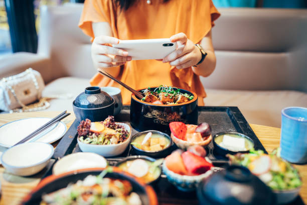 retrato da mulher asiática nova que toma o retrato de seu alimento japonês fresco com o telefone móvel no restaurante. - ethnic food - fotografias e filmes do acervo