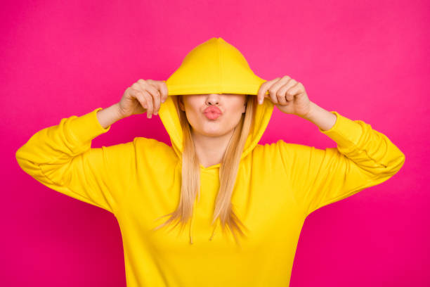 foto von netten dame versteckt augen mit kapuze senden luft kuss tragen gelb hoodie pullover isoliert rosa hintergrund - smiling women blond hair human face stock-fotos und bilder