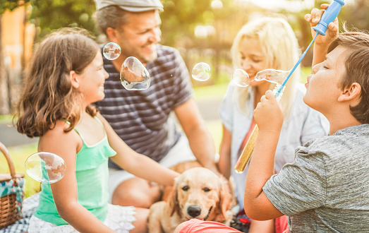 Happy family doing picnic in nature outdoor - Young parents having fun with children and their dog in summer time laughing, playing together with soap bubbles - Focus on boy eye