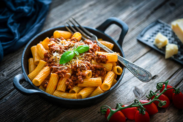Rigatoni pasta with Bolognese sauce shot on rustic wooden table Rigatoni Bolognese sauce pasta in a cast iron cooking pan shot on rustic wooden table. Some ingredients like ripe tomatoes, olive oil, basil, peppercorns and Parmesan cheese are all around the plate. XXXL 42Mp studio photo taken with SONY A7rII and Sony FE 90mm f2.8 Macro G OSS lens rigatoni stock pictures, royalty-free photos & images