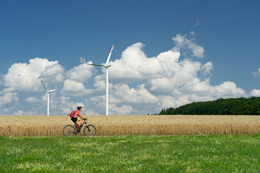 nice, active woman, riding her electric mountain bike between wheat fields and wind wheewls of a wind farm on the Swabian Alb near the city of Aalen, Baden-Wuerttemberg, Germany