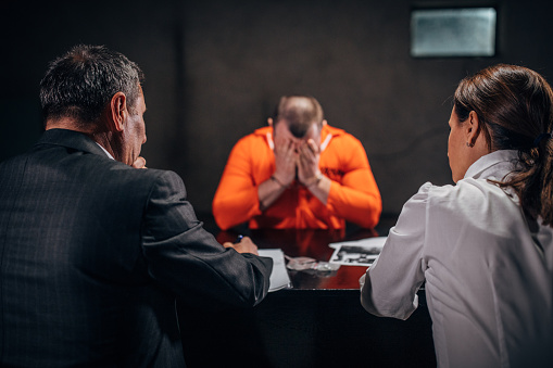 Three people, man and woman detectives interrogating a man prisoner in dark investigation room.