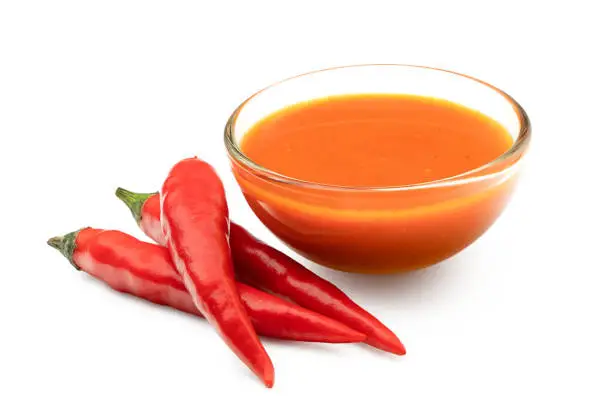 Photo of Peri peri chilli sauce in a glass bowl next to three red chillies isolated on white.