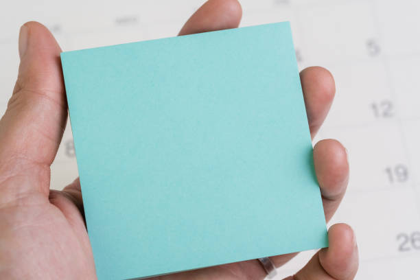 Hand holding blue sticky note or adhesive paper with copy space for writing message on white clean calendar using as reminder for vacation, project launch date or special event message Hand holding blue sticky note or adhesive paper with copy space for writing message on white clean calendar using as reminder for vacation, project launch date or special event message. calendar today personal organizer routine stock pictures, royalty-free photos & images