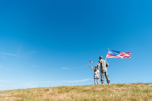 back view of kid in straw hat and military father holding american flags while standing on grass
