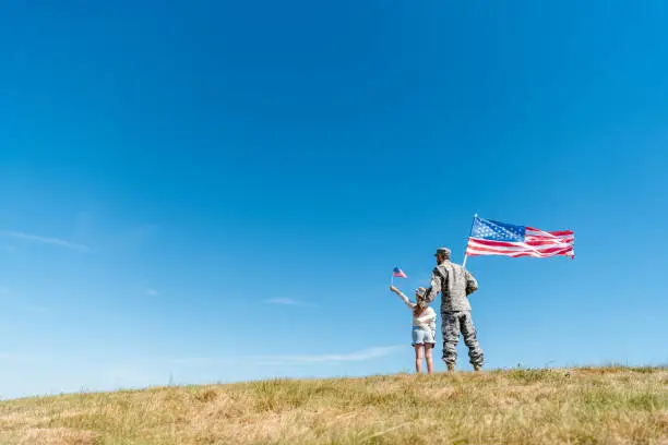 Photo of back view of kid in straw hat and military father holding american flags while standing on grass