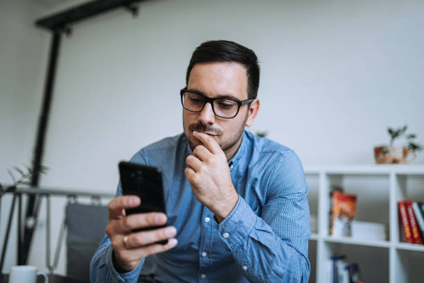 joven hombre reflexivo mirando el teléfono móvil. - incertidumbre fotografías e imágenes de stock