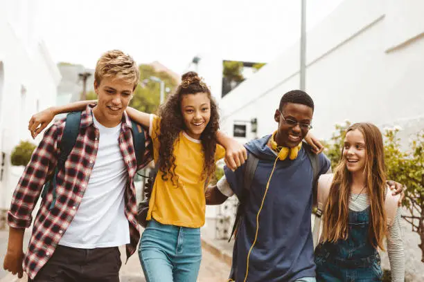 Smiling teenage friends wearing college bags having fun walking together in the street. Happy teenage boys and girls walking in an alley holding each other.