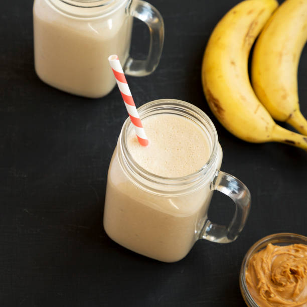 Mason glass jar mugs filled with peanut butter banana smoothie on a black background, low angle view.