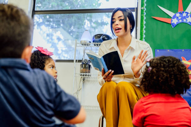 Hispanic schoolteacher reading aloud to her young students Young female Hispanic schoolteacher reading aloud to elementary aged students sitting around her on the floor. elementary age stock pictures, royalty-free photos & images