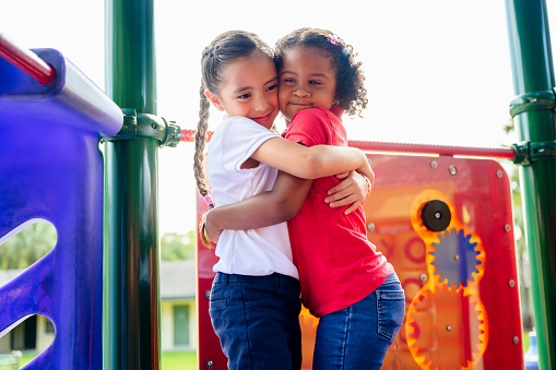 Affectionate young female Hispanic best friends hugging outdoors on playground equipment during school recess break.