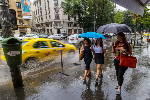 Bucharest, Romania - August 01, 2019: Women walking with umbrellas, in rainy weather, while a taxi splashes them with water from a pond in downtown Bucharest. This image is for editorial use only.