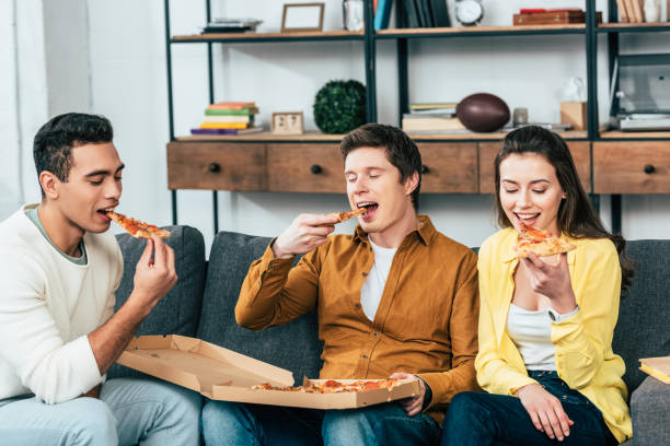 istock Three multiethnic joyful friends sitting on sofa and eating pizza together 1166254821