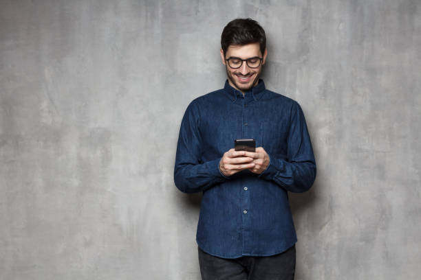 istock Young smiling man browsing internet or typing a message using phone, standing against gray textured wall with copy space 1166255853