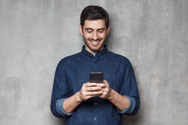 uomo d'affari smiiling in camicia di jeans e occhiali alla moda in piedi contro la parete grigia strutturata, digitando un messaggio utilizzando il suo telefono - smart casual foto e immagini stock