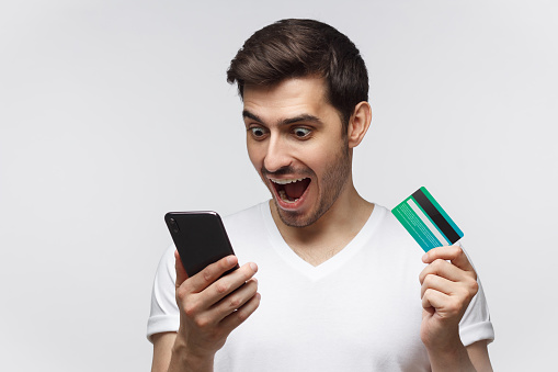 Portrait of excited young handsome man with mouth open, holding credit card, using mobile phone, isolated on gray background