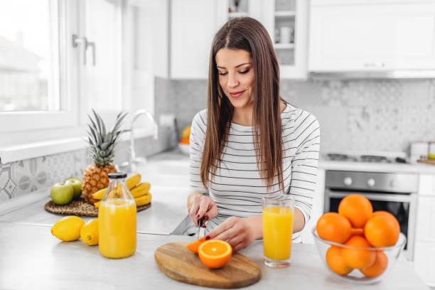 Smiling young woman in a gray T-shirt squeezes a fresh orange on fruit juice. Healthy eating concept. In the interior of the kitchen stock photo