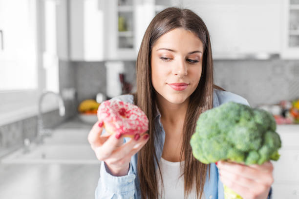 Young woman in blue shirt choosing between broccoli or junk food, doughnut. Healthy clean detox eating concept. Vegetarian, vegan, raw concept. Copy space. stock photo