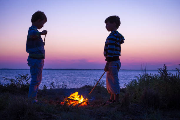 two kids on the beach with campfire at sunset - bonfire beach fire barbecue imagens e fotografias de stock