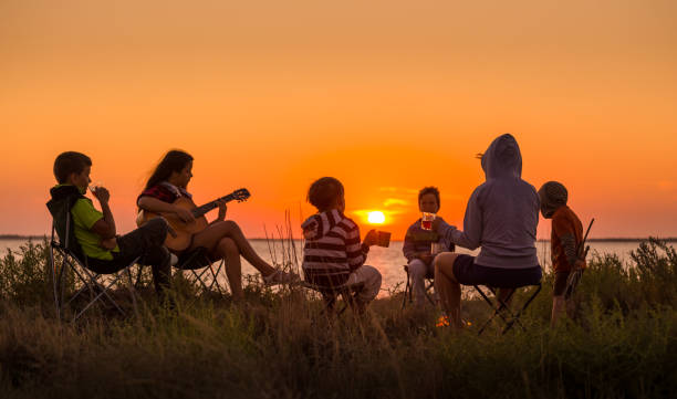 les gens s'asseyant sur la plage avec le feu de camp au coucher du soleil - camping friendship campfire fire photos et images de collection