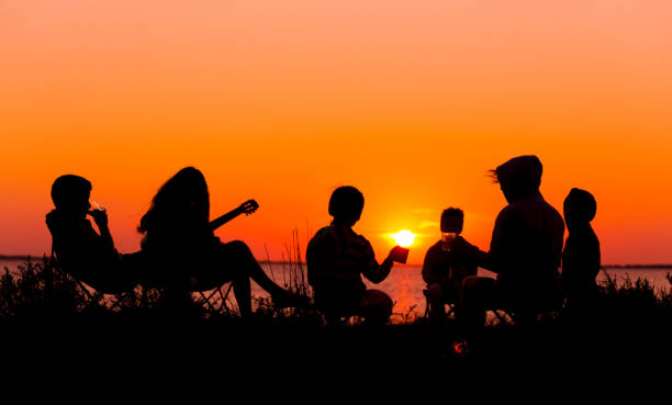 silhouette of people sitting on the beach with campfire at sunse - picnic family barbecue social gathering imagens e fotografias de stock