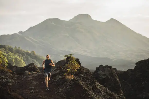 Photo of Young athlete man trail running in mountains in the morning. Amazing volcanic landscape of Bali mount Batur on background. Healthy lifestyle concept.