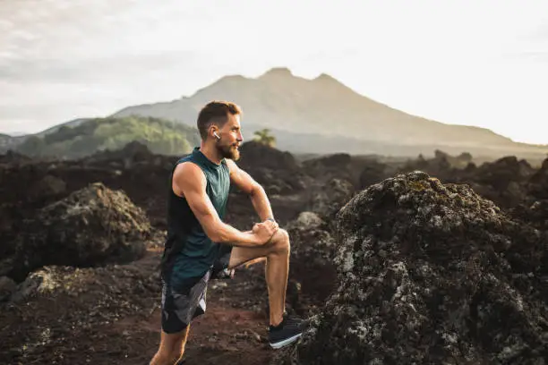 Photo of Young hipster runner with beard stretching and warming-up for trail running outdoors. Listening music in air pods. Mountain view on background.