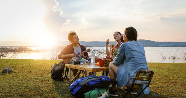 un gruppo di amici asiatici turisti che bevono e suonano la chitarra insieme alla felicità in estate mentre si accampano vicino al lago al tramonto - friendship camping night campfire foto e immagini stock