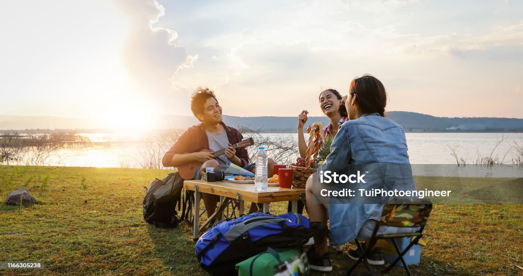 A group of Asian friends tourist drinking and playing guitar together with happiness in Summer while having camping near lake at sunset Friendship Stock Photo