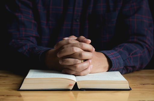 Hands of a young man folded praying over a Bible on wooden table