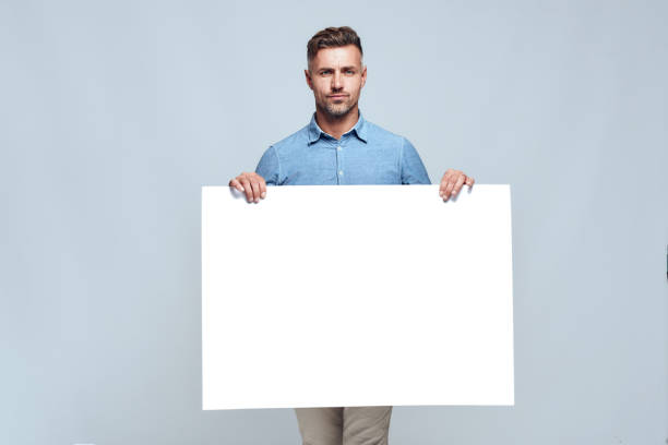 tu texto aquí. retrato de hombre barbudo guapo en ropa casual sosteniendo tablero vacío en blanco y mirando la cámara mientras está de pie contra fondo gris - man holding a sign fotografías e imágenes de stock