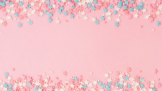 Festive border frame of colorful pastel sprinkles on pink background with copy space in center. Sugar sprinkle dots and stars, decoration for cake and bakery. Top view or flat lay. Banner