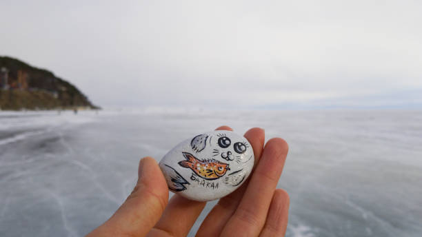 Female hand fingers holding a small decorative magnet with the inscription in Cyrillic "Baikal". Souvenir from Siberia. On the background of mountains and ice lake Baikal stock photo